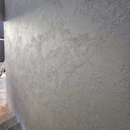 HYDBUD - Szara ściana z dekoracyjną, nierównomierną fakturą, widoczna z bliska, z fragmentem schodów w dolnym lewym rogu.