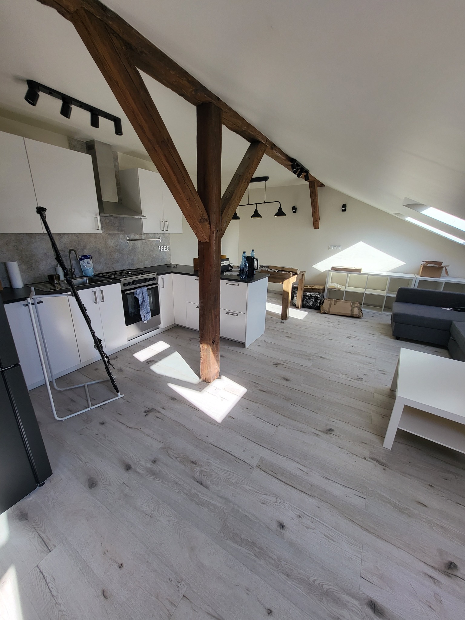 Jasne poddasze z drewnianą konstrukcją nośną, białą zabudową kuchenną i panelami podłogowymi w odcieniach szarości, częściowo umeblowane, z wpadającym przez okno dachowe światłem słonecznym.