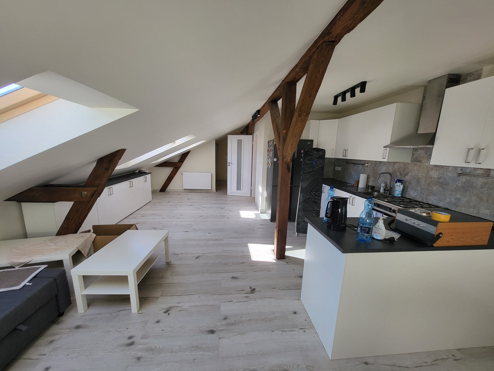 Jasne, przestronne poddasze z drewnianymi belkami stropowymi, białą zabudową kuchenną, wyspą i panelami podłogowymi w odcieniach szarości; widok na otwartą przestrzeń z oknami dachowymi.