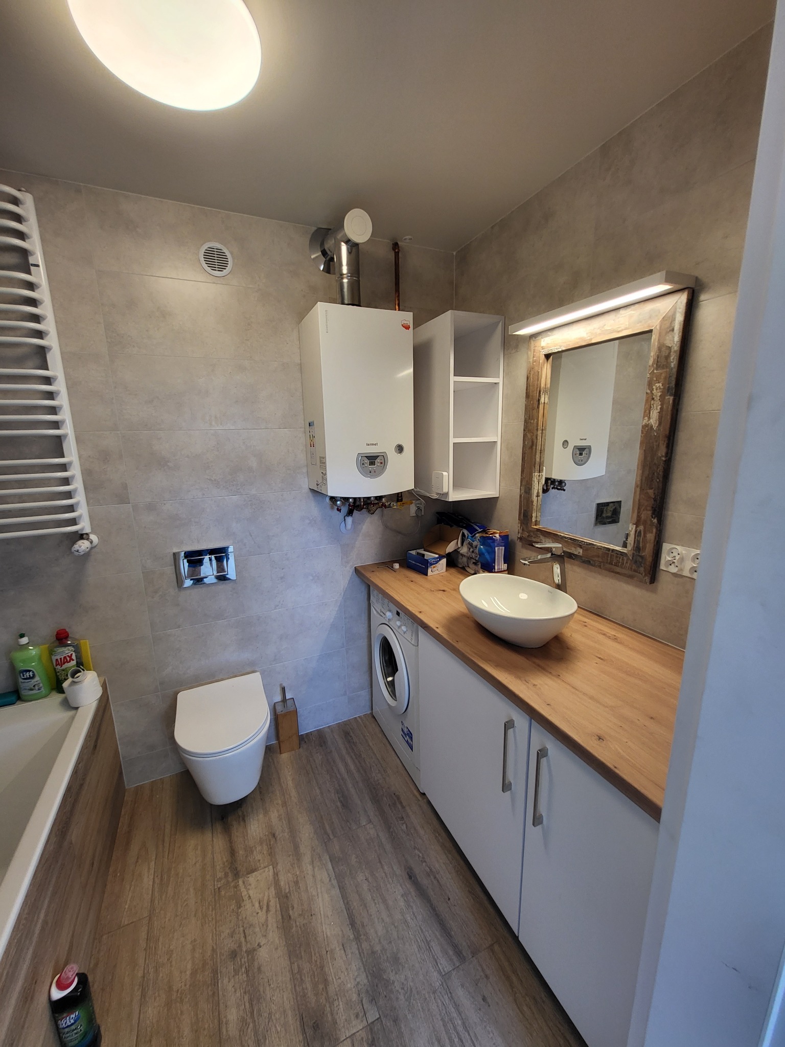 Wykończona łazienka w Obornikach Śląskich z drewnopodobnymi płytkami podłogowymi, umywalką nablatową, szafką pod umywalką, lustrem w ozdobnej ramie i grzejnikiem drabinkowym.