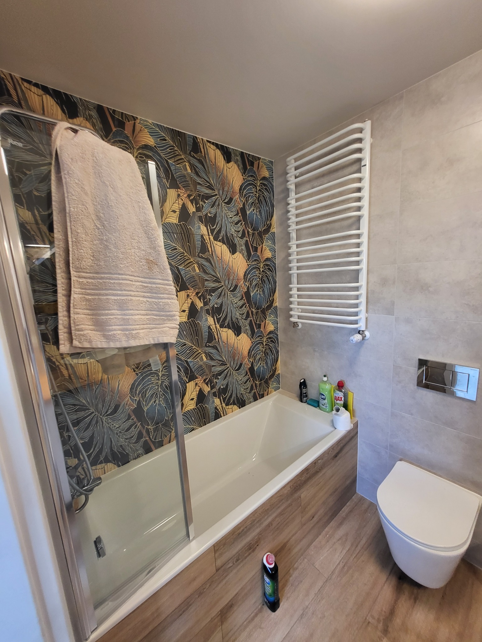 Nowoczesna łazienka z wanną za szklanym parawanem, egzotyczną tapetą w liście i grzejnikiem drabinkowym, widok na wiszącą toaletę i butelkę płynu do czyszczenia na podłodze.