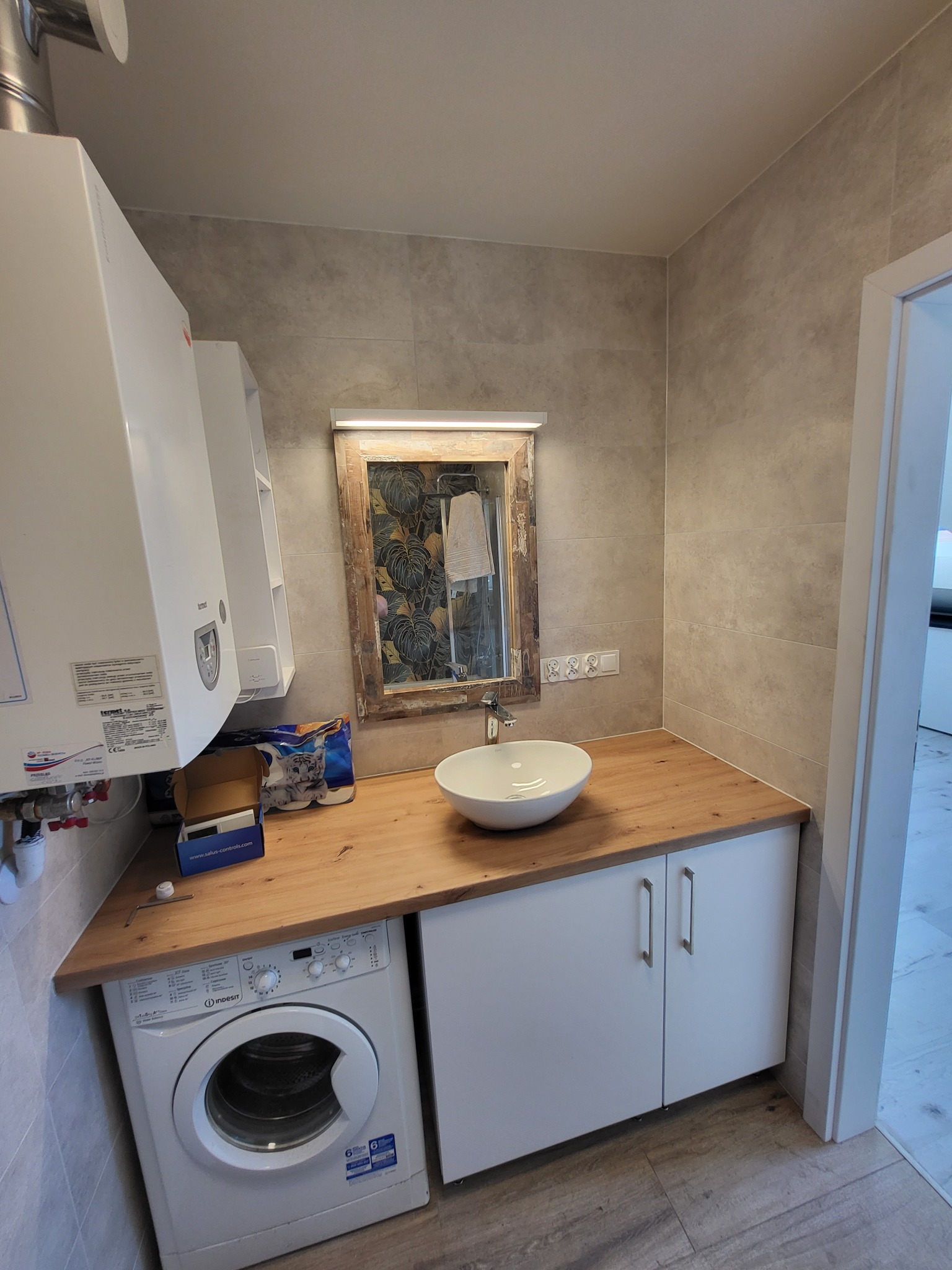 Wykończona łazienka w beżowych płytkach z drewnianym blatem, białą umywalką nablatową, szafką pod umywalką, pralką Indesit i lustrem w ozdobnej ramie z podświetleniem LED.