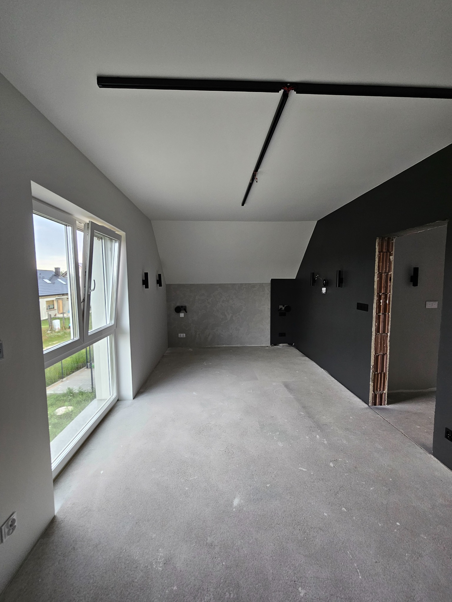 Surowe wnętrze pokoju z otwartym oknem, szarą wylewką, czarną ścianą z wyprowadzonymi kablami i widoczną konstrukcją ceglaną otworu drzwiowego.