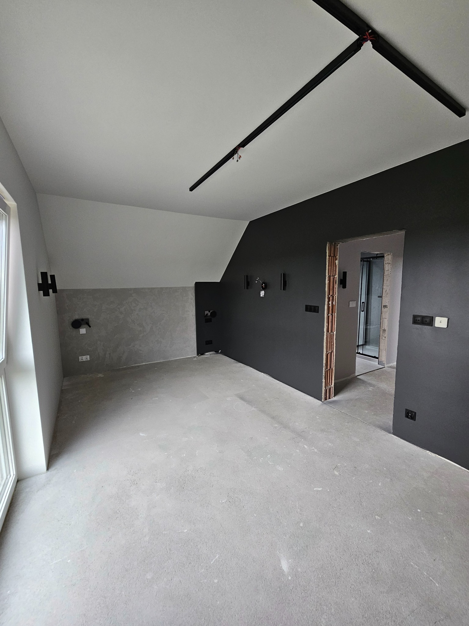 Wnętrze pokoju w trakcie remontu z surową podłogą, pomalowanymi na szaro ścianami z widocznymi instalacjami elektrycznymi, czarną listwą oświetleniową na suficie i otwartym przejściem do łazienki...