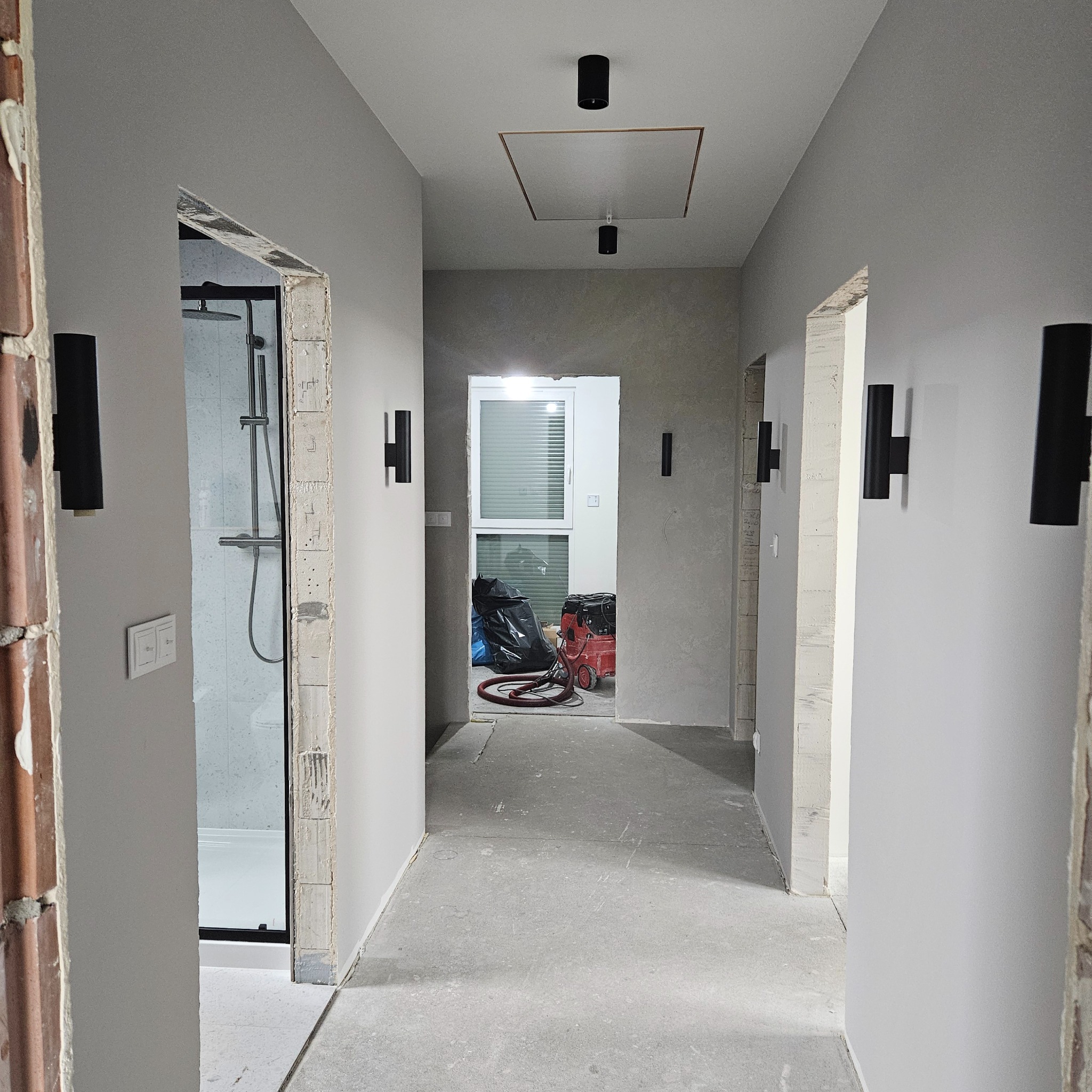 Korytarz w trakcie prac wykończeniowych: surowe ściany pomalowane na szaro, widoczne czarne lampy ścienne, niezabezpieczone ościeżnice drzwiowe, w tle widoczny remontowany pokój z workami na śmieci...