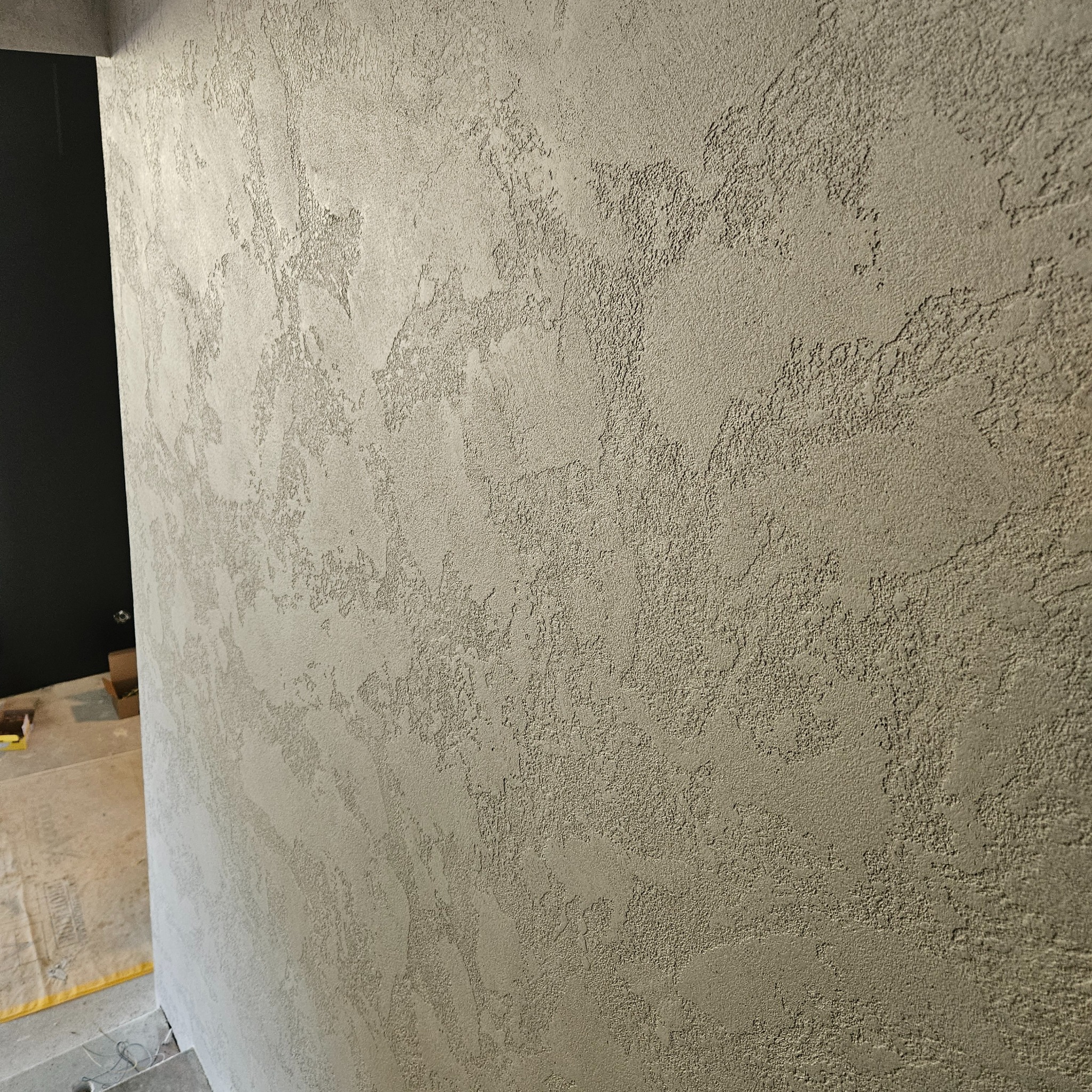 Ściana pokryta dekoracyjnym tynkiem strukturalnym w odcieniach szarości, z widoczną fakturą i nieregularnym wzorem, w tle fragment czarnego otworu drzwiowego i zabezpieczona podłoga.