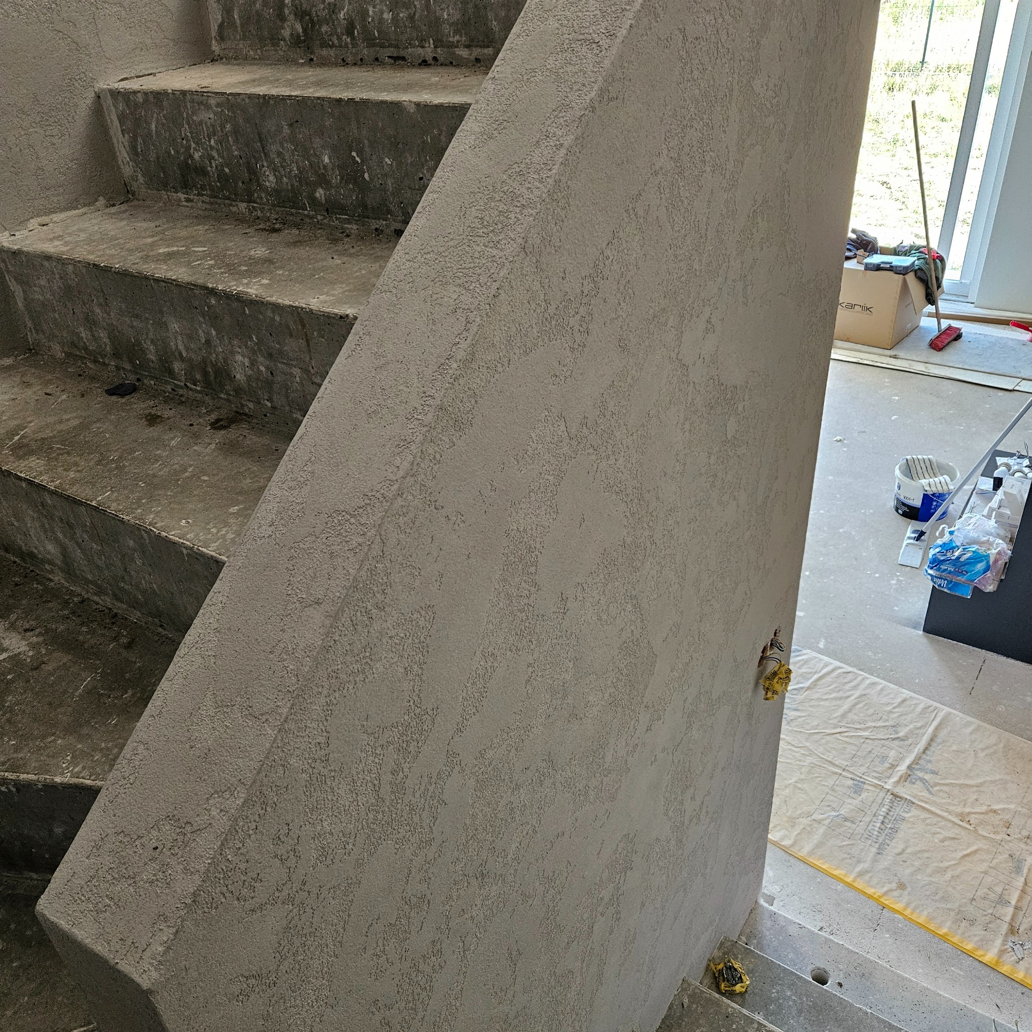 Surowe betonowe schody w trakcie wykańczania, widoczna ściana z nałożonym tynkiem strukturalnym, zabezpieczona podłoga, wystające przewody elektryczne.