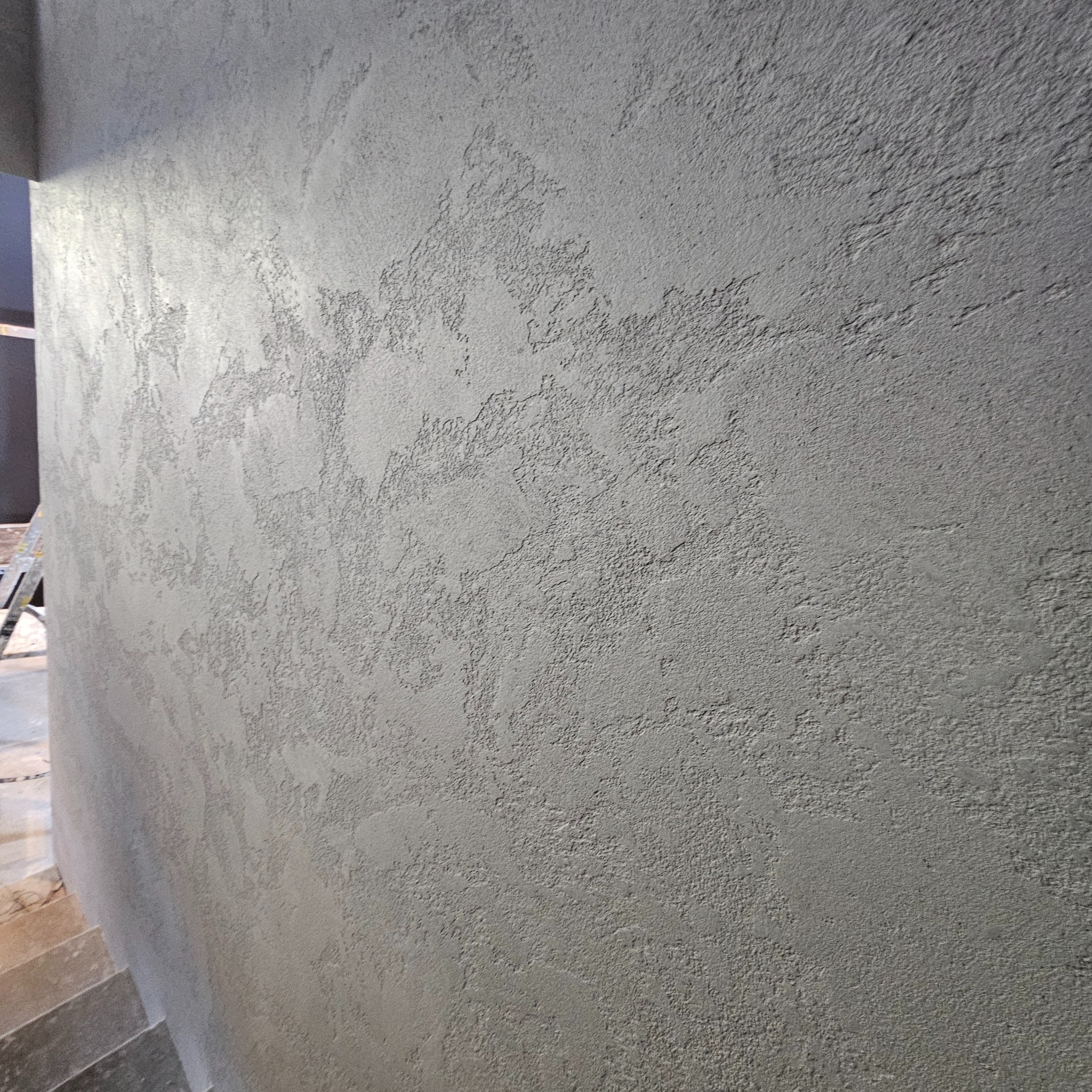 Szara ściana z dekoracyjną, nierównomierną fakturą, widoczna z bliska, z fragmentem schodów w dolnym lewym rogu.