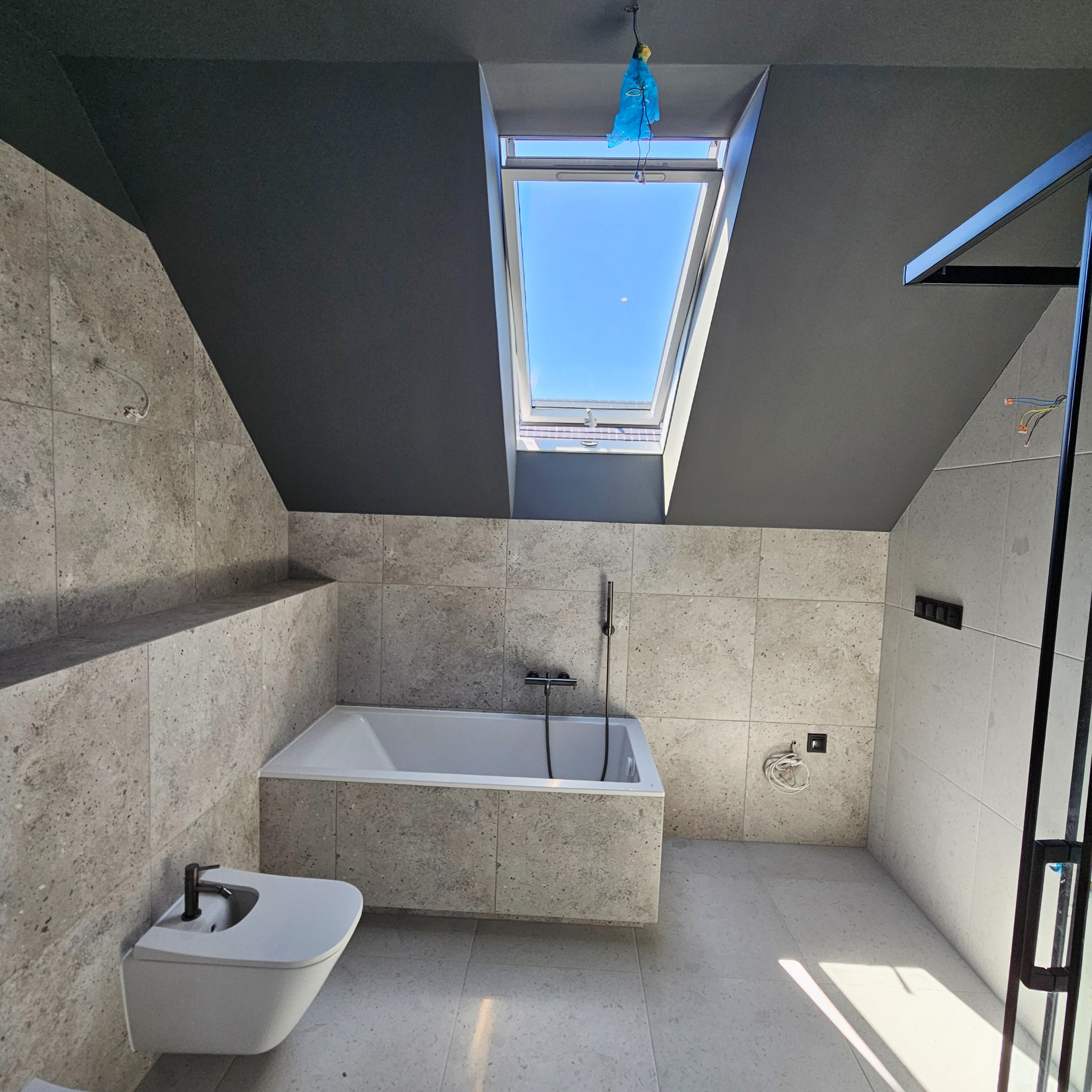 Nowoczesna łazienka na poddaszu z oknem dachowym, wykończona płytkami imitującymi kamień w odcieniach szarości, z białą wanną, bidetem i czarnym sufitem. Widoczne wystające przewody elektryczne...