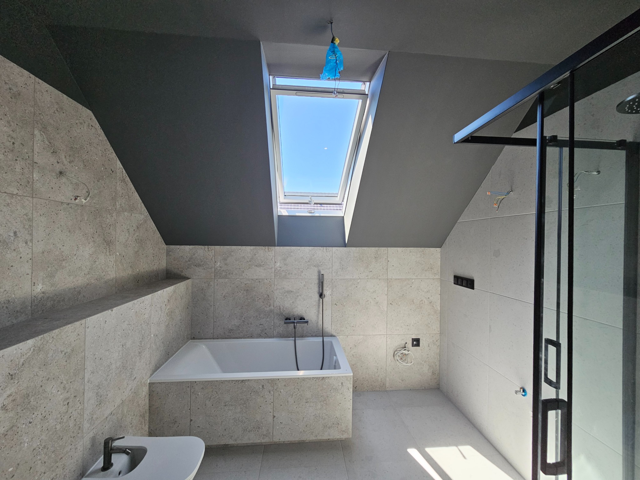 Nowoczesna łazienka na poddaszu z oknem dachowym, wykończona płytkami w odcieniach szarości, z wanną, kabiną prysznicową z czarnymi ramami i ciemnoszarym sufitem.