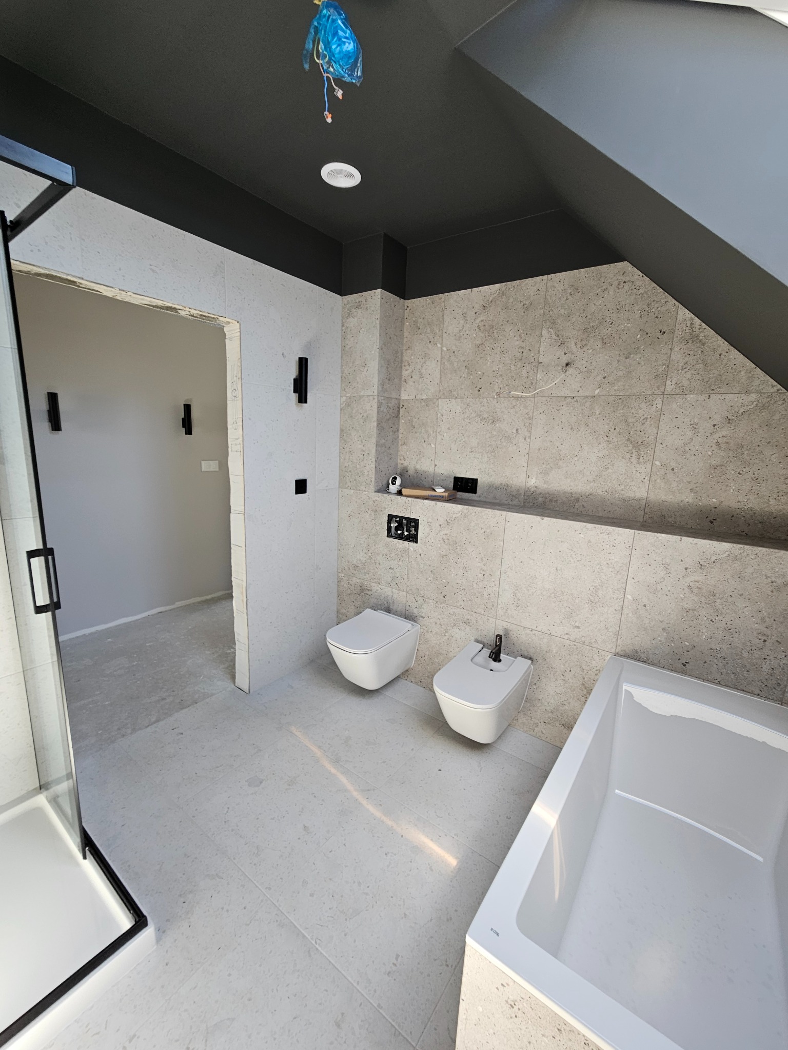 Wnętrze łazienki w trakcie wykańczania, widoczna wanna, toaleta i bidet w zabudowie, ściany wyłożone płytkami imitującymi kamień, czarny sufit pod skosem, instalacja elektryczna zabezpieczona folią.