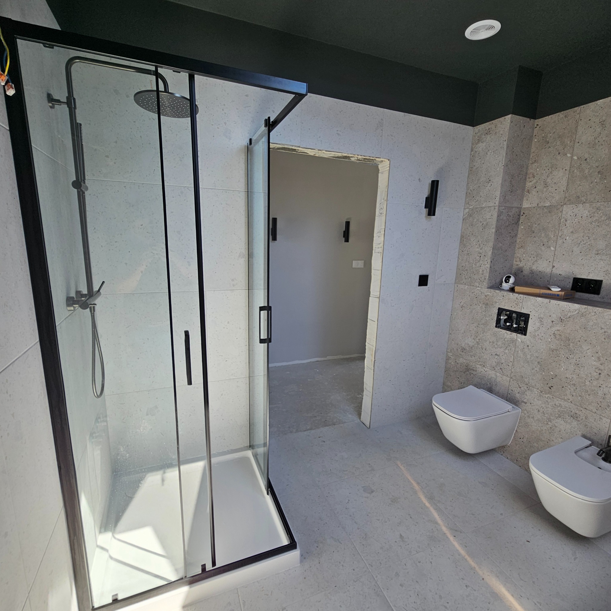 Nowoczesna łazienka z kabiną prysznicową w czarnej ramie, wiszącą toaletą i bidetem, szare płytki na ścianach i podłodze, czarny sufit, drzwi do innego pomieszczenia w tle.