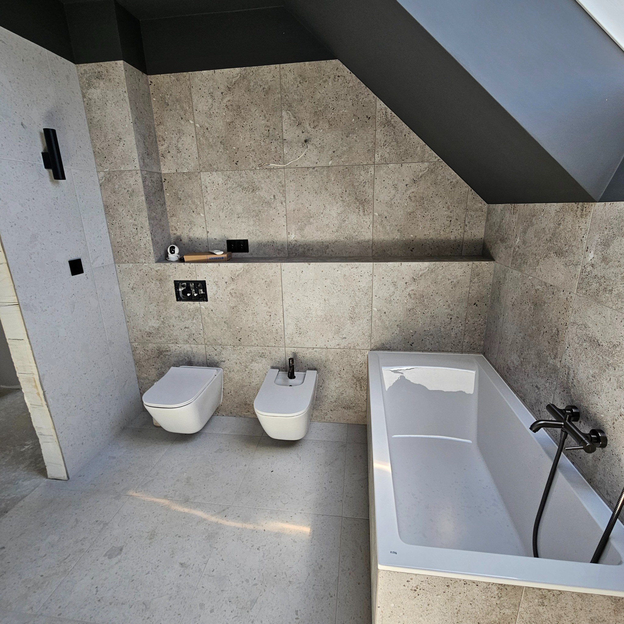Nowoczesna łazienka w szarościach z wanną, toaletą i bidetem, wykończona płytkami imitującymi kamień, z czarną armaturą i oświetleniem.