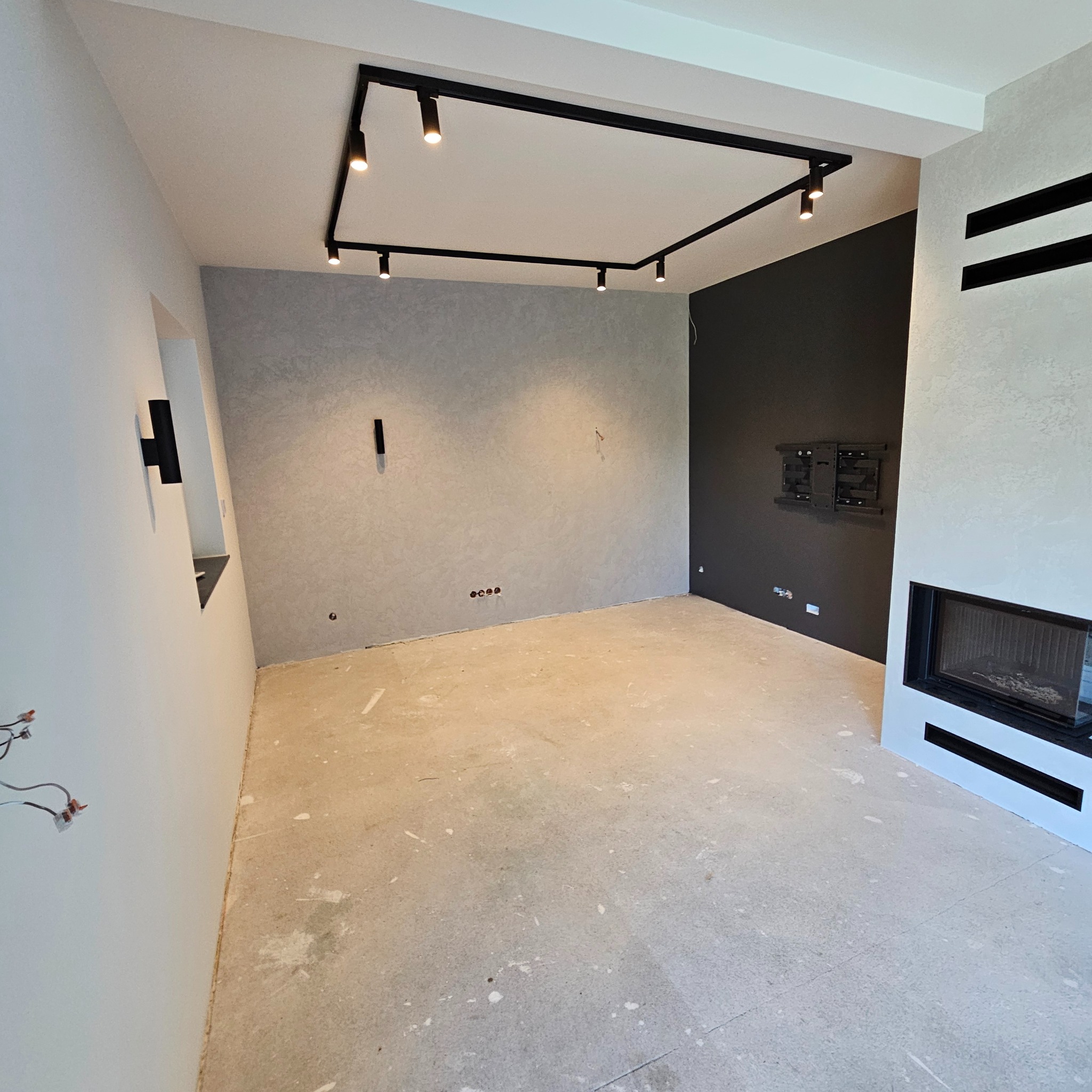 Wnętrze pokoju po pracach wykończeniowych, widoczne ściany w odcieniach szarości i czerni, sufit z nowoczesnym oświetleniem szynowym, przygotowane miejsce na telewizor oraz kominek z czarnymi...