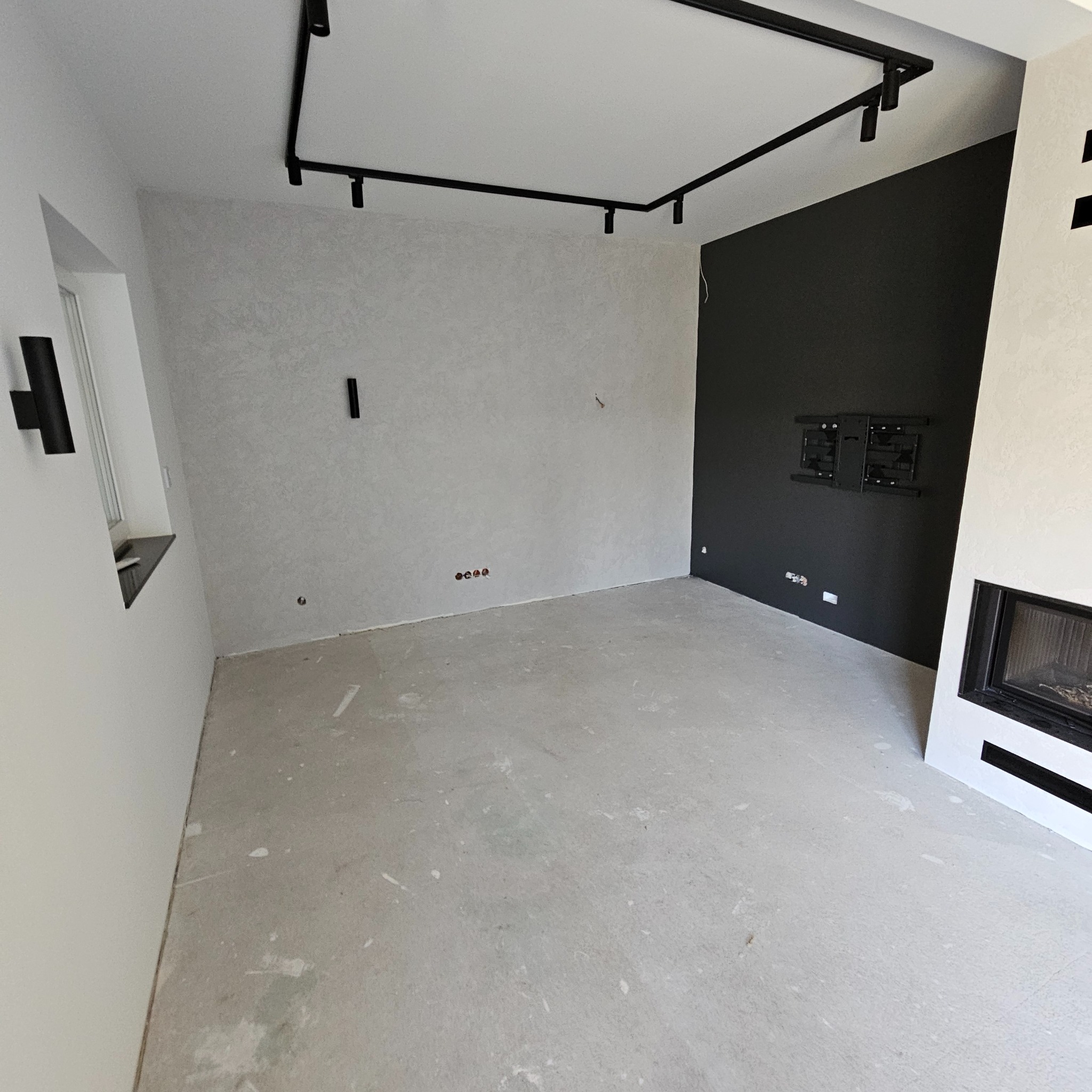 Wnętrze pokoju po remoncie z czarną ścianą, zamontowanym uchwytem pod telewizor, kominkiem i szynoprzewodem oświetleniowym.