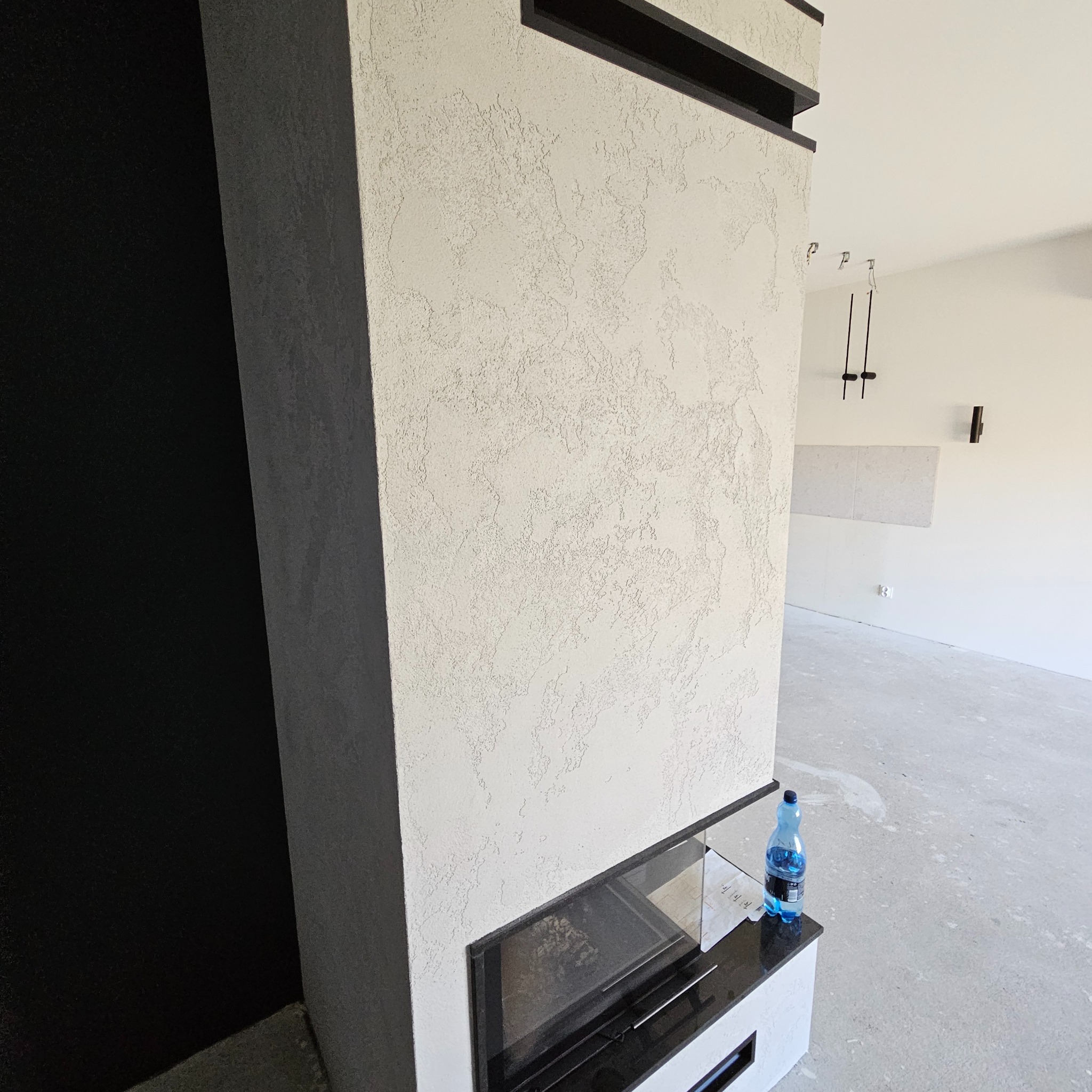Fragment ściany z dekoracyjnym, jasnym tynkiem strukturalnym przy czarnym obramowaniu kominka; na kominku stoi butelka wody.