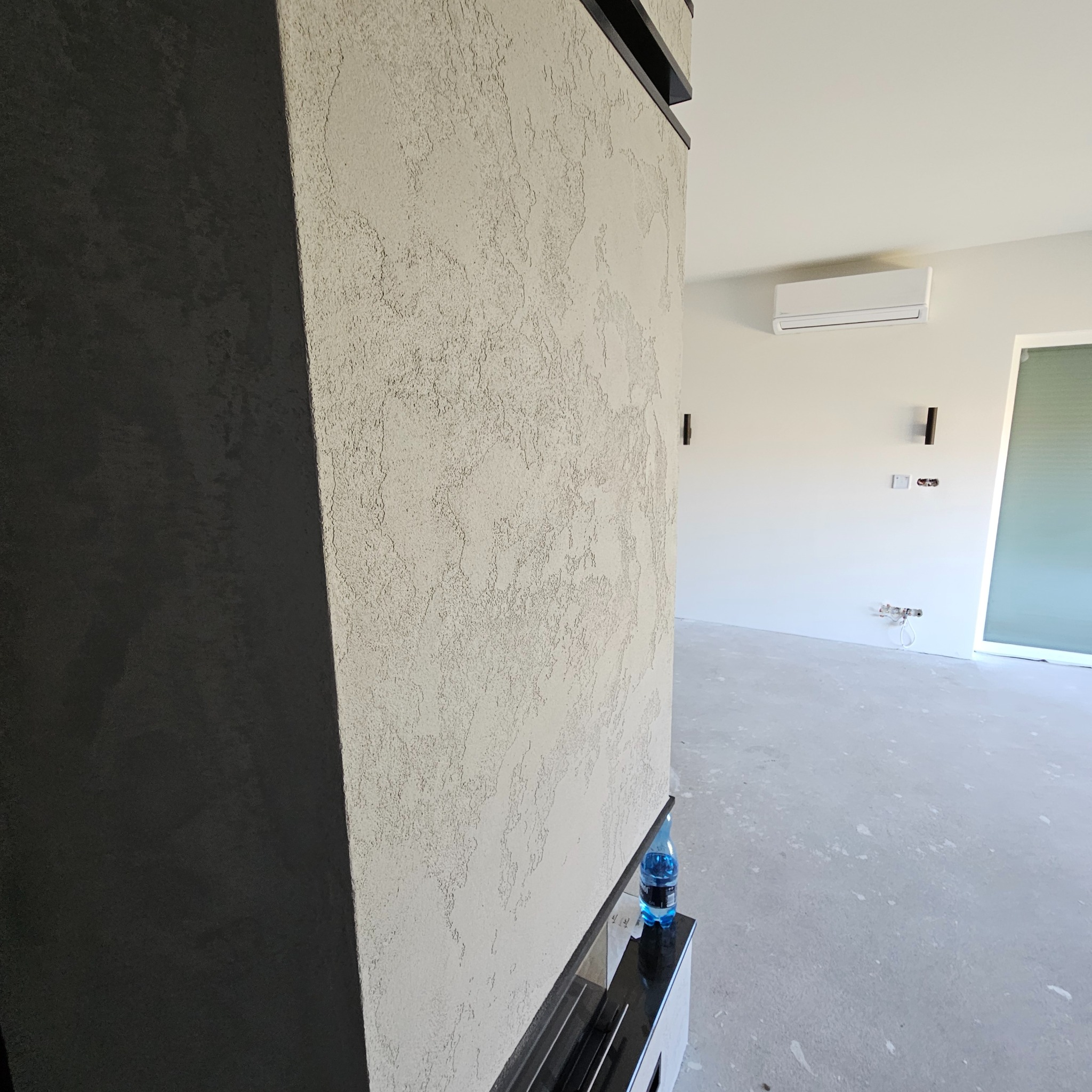 Fragment ściany z dekoracyjną strukturą tynku w odcieniach beżu, obok czarny element konstrukcyjny, w tle jasne pomieszczenie z klimatyzatorem i wystającymi przewodami instalacji elektrycznej...