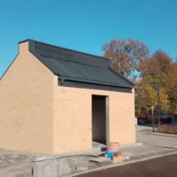 Nowy, blaszany dach z obróbkami na małym, murowanym budynku gospodarczym z cegły klinkierowej, na tle jesiennego krajobrazu i błękitnego nieba.