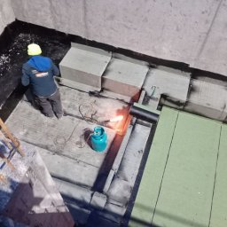 SIM DIM House - Prace dekarskie: Widok z góry na dekarza w kasku, z palnikiem gazowym i butlą, podczas układania papy termozgrzewalnej na betonowym podłożu, widoczne fragmenty izolacji i obróbki blacharskiej.