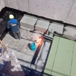 Prace dekarskie: Widok z góry na dekarza w kasku, z palnikiem gazowym i butlą, podczas układania papy termozgrzewalnej na betonowym podłożu, widoczne fragmenty izolacji i obróbki blacharskiej.