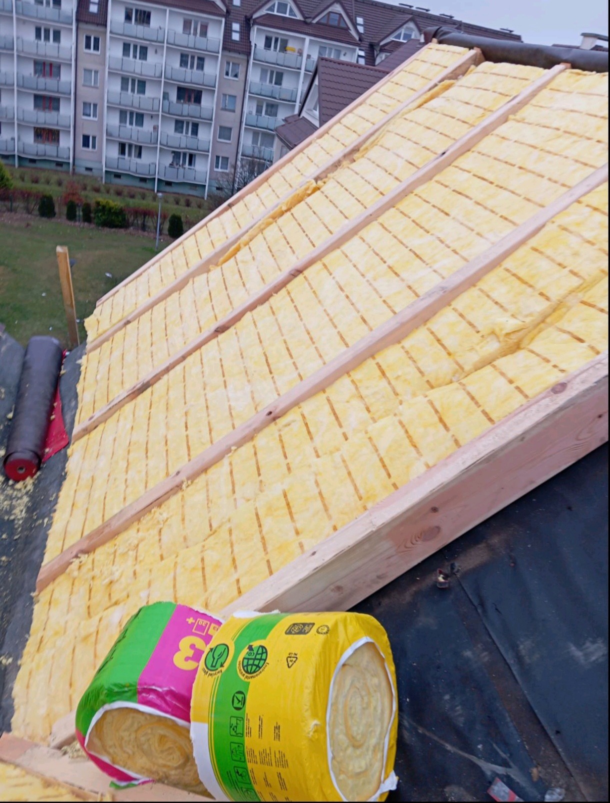 Poddasze w trakcie ocieplania wełną mineralną, widoczne rolki wełny w kolorowych opakowaniach, konstrukcja dachu z łatami, w tle budynek mieszkalny.