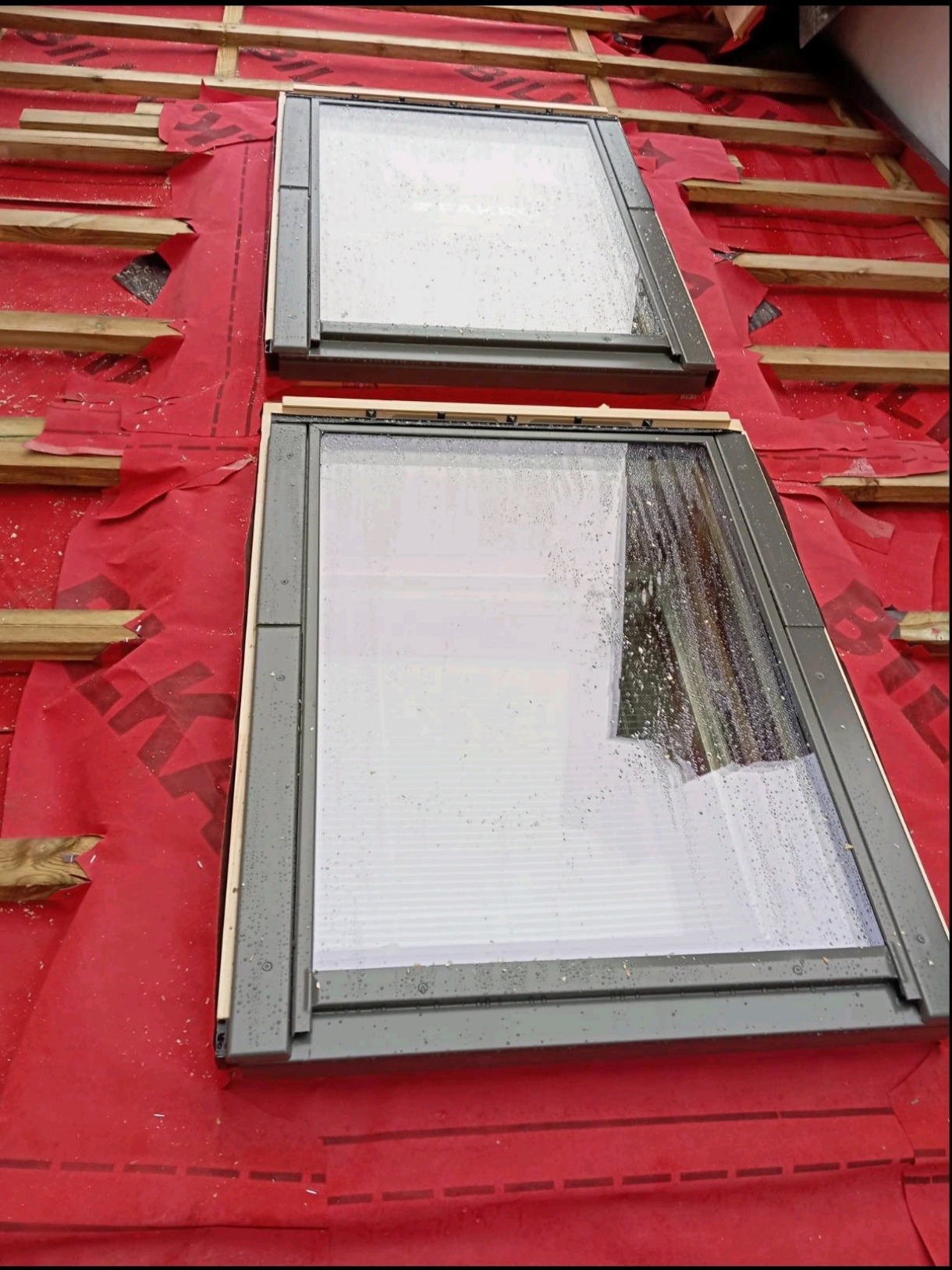 Dwa okna dachowe z szarą ramą, osadzone w czerwonej membranie dachowej, widoczne drewniane elementy konstrukcji dachu.