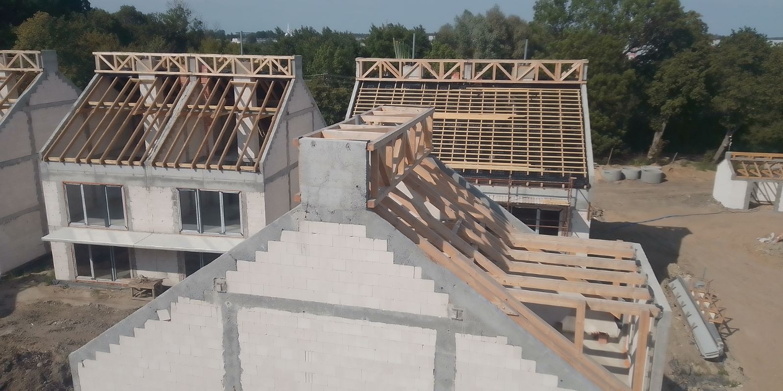 Budowa domów w stanie surowym otwartym, widoczne krokwie dachowe, komin z betonu komórkowego i pustaków ceramicznych, teren budowy z drzewami w tle.