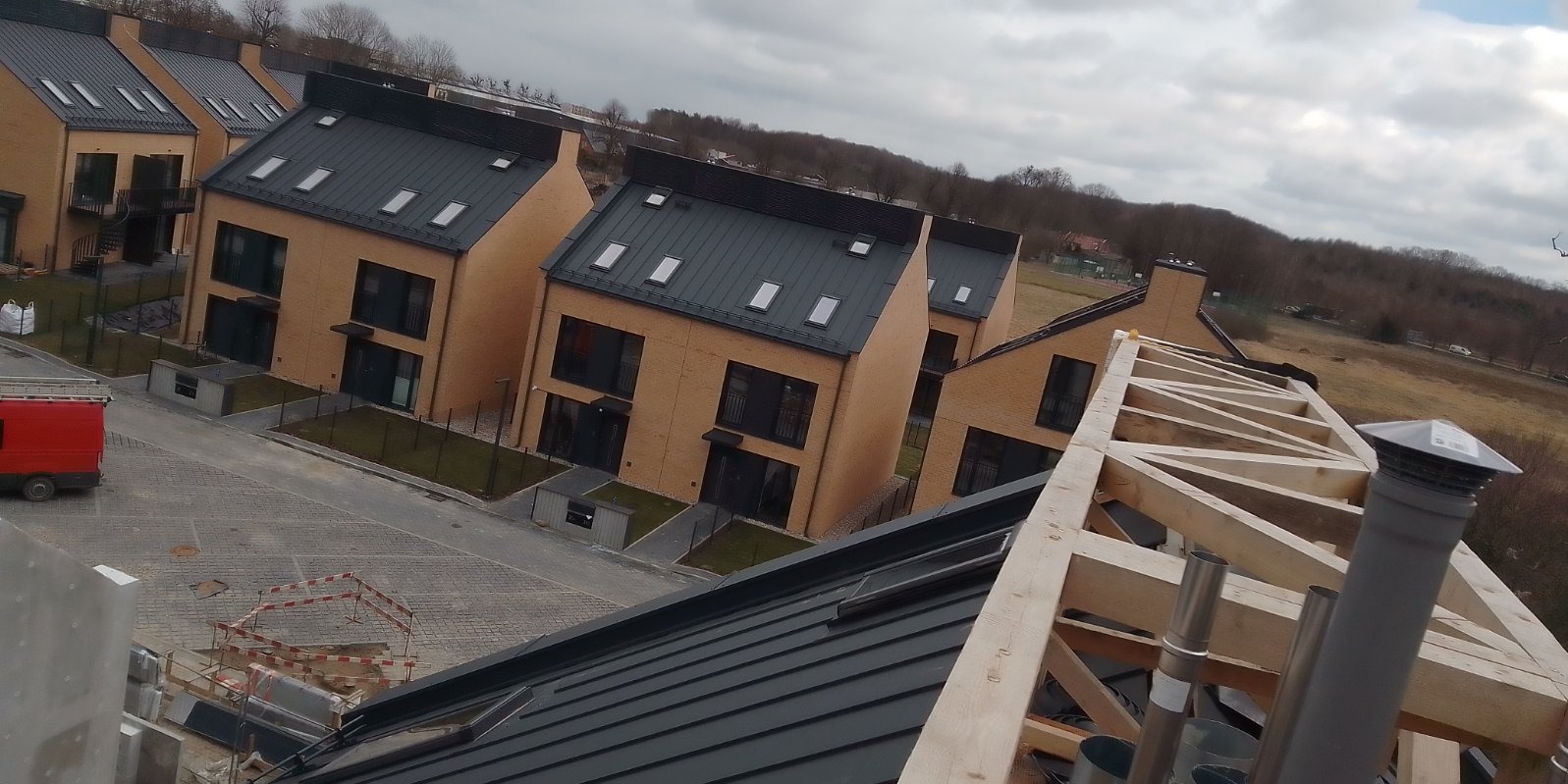 Widok z góry na konstrukcję drewnianą dachu w trakcie budowy, z widocznymi nowymi domami w tle i elementami wentylacji kominowej.
