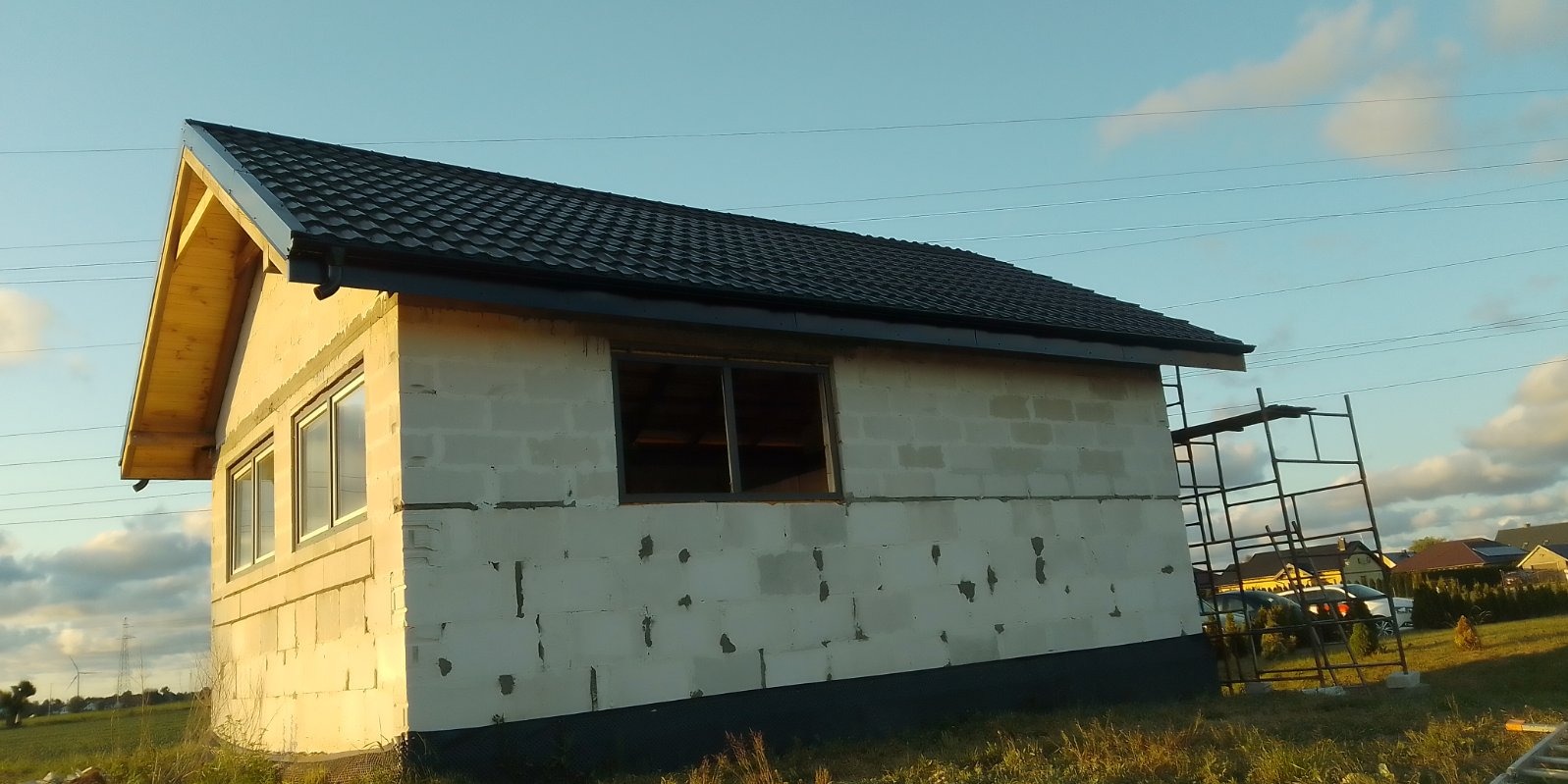 Niedokończony budynek w trakcie budowy, z czarnym dachem z blachodachówki, oknami bez szyb, oraz rusztowaniem z boku, na tle błękitnego nieba i zielonego krajobrazu.