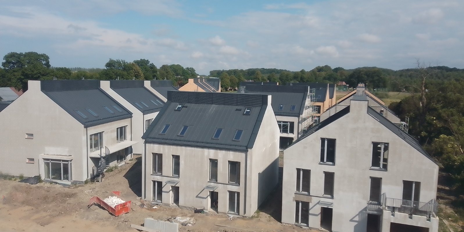 Nowe domy szeregowe z ciemnymi dachami i oknami dachowymi, widoczne prace wykończeniowe na zewnątrz, gruz w czerwonym kontenerze.