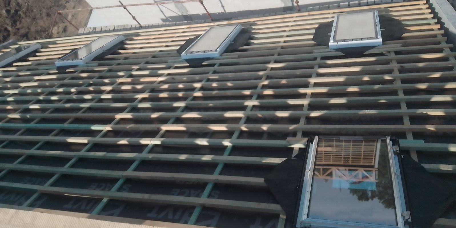 Montaż okien dachowych na drewnianej konstrukcji dachu, widoczna membrana dachowa i rusztowanie w tle.