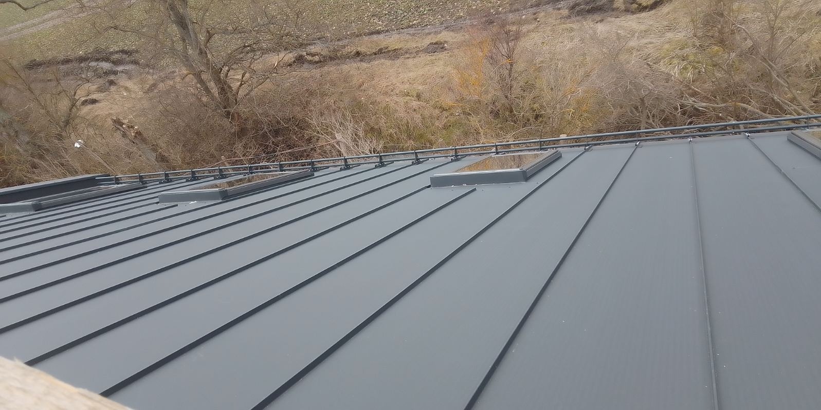 Szary dach z blachy trapezowej z trzema oknami dachowymi, zabezpieczony barierką śniegową, na tle krajobrazu z drzewami i wzgórzami.