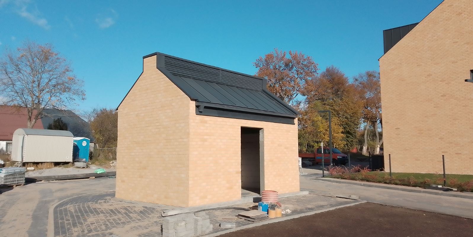 Nowy, blaszany dach z obróbkami na małym, murowanym budynku gospodarczym z cegły klinkierowej, na tle jesiennego krajobrazu i błękitnego nieba.