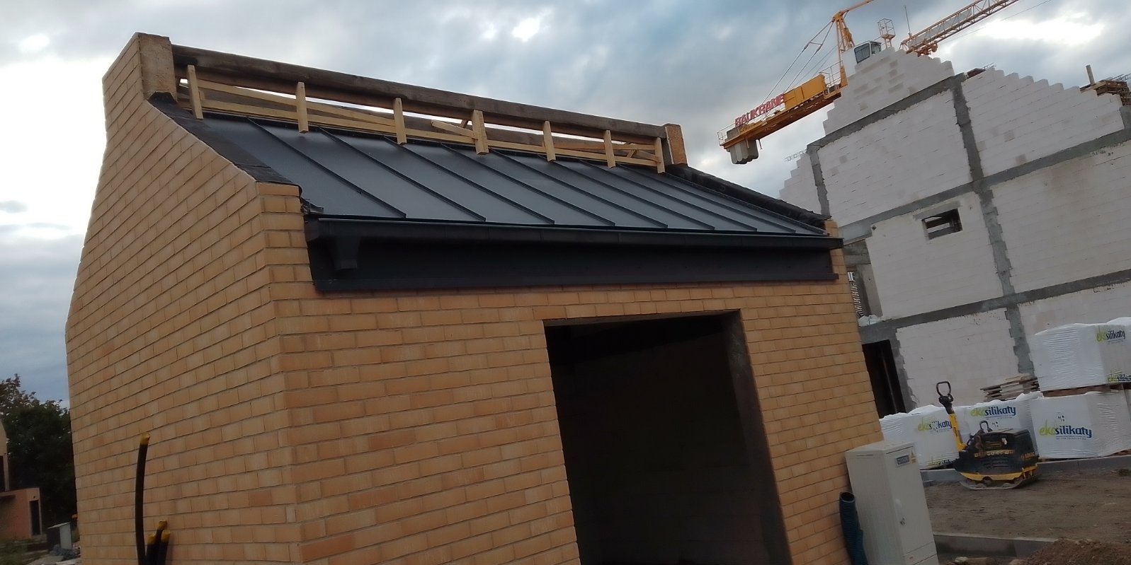 Budynek z cegły z nowym, czarnym, metalowym pokryciem dachowym, drewnianą konstrukcją pod przyszły dach, widoczne obróbki blacharskie, w tle budowa i dźwig.