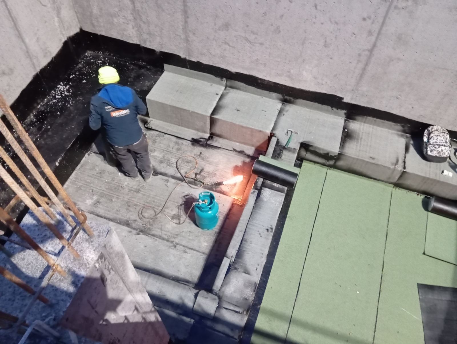 Prace dekarskie: Widok z góry na dekarza w kasku, z palnikiem gazowym i butlą, podczas układania papy termozgrzewalnej na betonowym podłożu, widoczne fragmenty izolacji i obróbki blacharskiej.