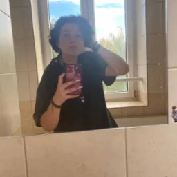 Osoba robiąca selfie w łazience, widoczne kafelki, okno z widokiem na drzewa i niebieskie niebo.