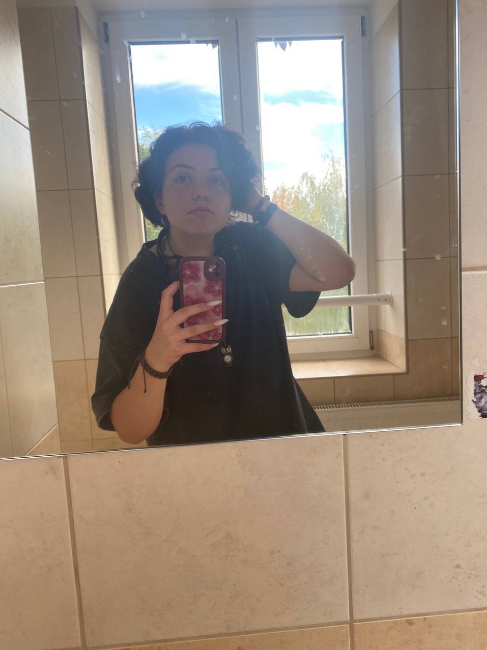 Osoba robiąca selfie w łazience, widoczne kafelki, okno z widokiem na drzewa i niebieskie niebo.