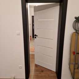 EMPEDE - Meble Podłogi Drzwi - Otwarta biała płycinowa drzwi wewnętrzne z czarną ościeżnicą na tle drewnianej podłogi i białej ściany z widocznym gniazdkiem elektrycznym i kubkiem przy podłodze.