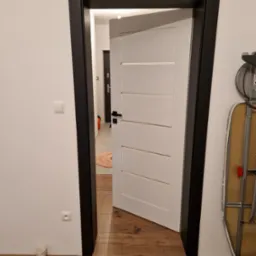 Otwarta biała płycinowa drzwi wewnętrzne z czarną ościeżnicą na tle drewnianej podłogi i białej ściany z widocznym gniazdkiem elektrycznym i kubkiem przy podłodze.