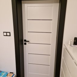 EMPEDE - Meble Podłogi Drzwi - Białe drzwi wewnętrzne z poziomymi frezowaniami i czarną ościeżnicą, widoczny fragment pokoju z komodą i paczką pieluch.