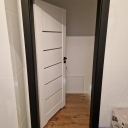 EMPEDE - Meble Podłogi Drzwi - Białe drzwi wewnętrzne z czarną ościeżnicą, częściowo otwarte, ukazujące drewnianą podłogę w pomieszczeniu za nimi; widoczne białe płytki na ścianie i pudełko z akcesoriami w rogu.