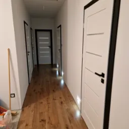 Korytarz z panelami podłogowymi w odcieniach brązu, białe drzwi z czarnymi ościeżnicami i klamkami, widoczne narzędzia i materiały budowlane przy ścianie.