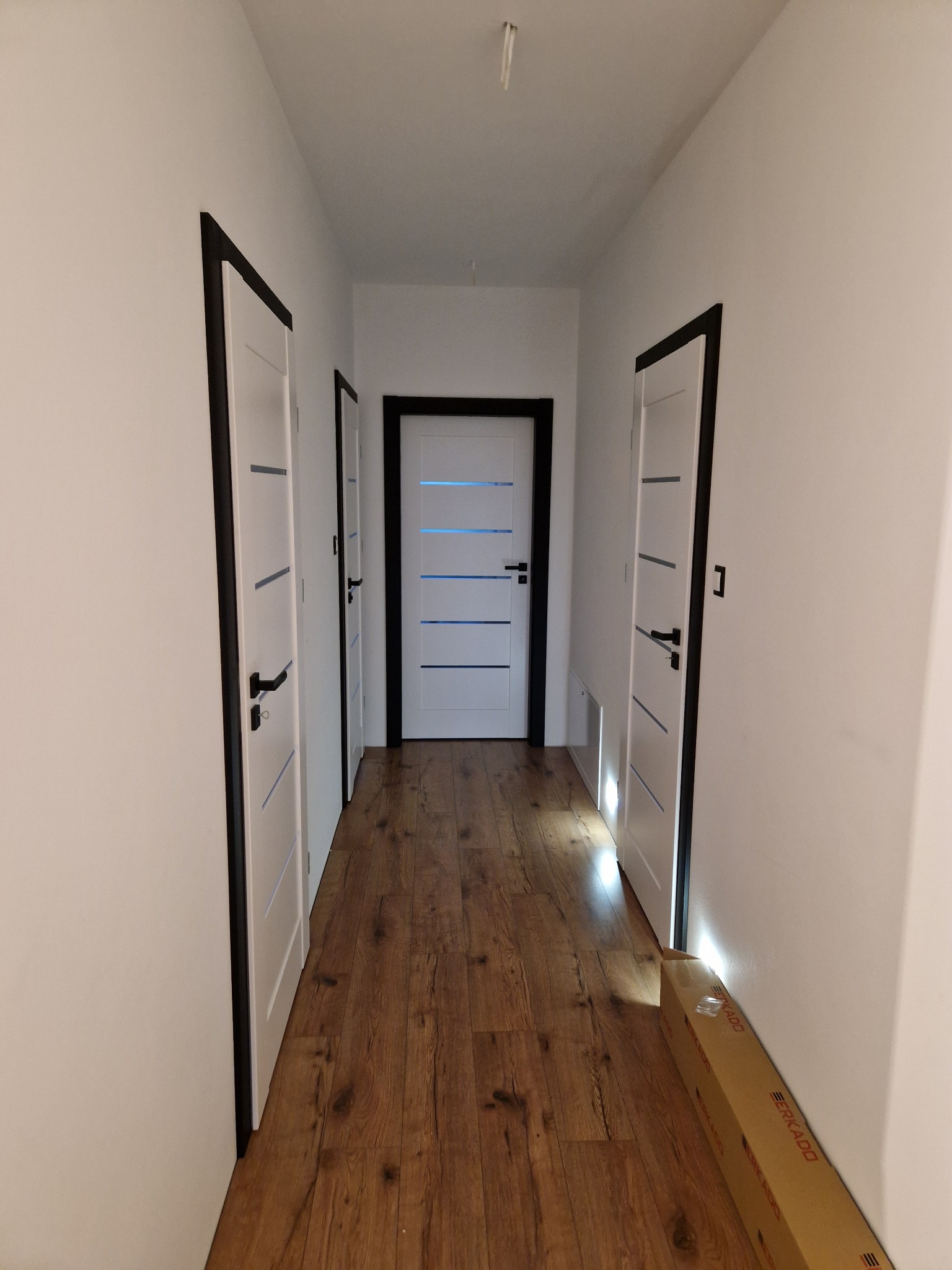Korytarz z trzema białymi drzwiami wewnętrznymi z czarnymi ościeżnicami i poziomymi przeszkleniami, brązowe panele podłogowe, kartonowe opakowanie przy ścianie.