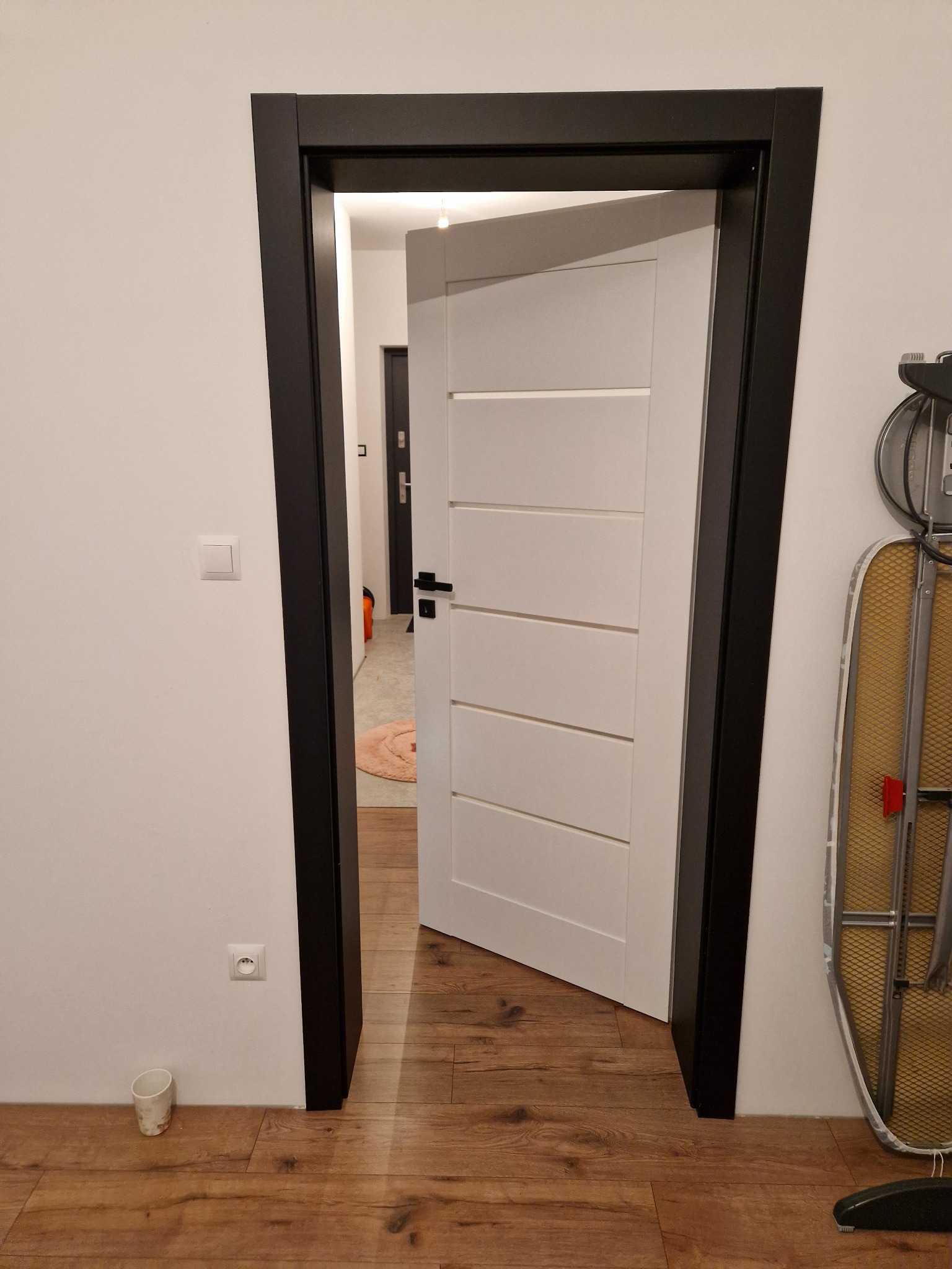 Otwarta biała płycinowa drzwi wewnętrzne z czarną ościeżnicą na tle drewnianej podłogi i białej ściany z widocznym gniazdkiem elektrycznym i kubkiem przy podłodze.