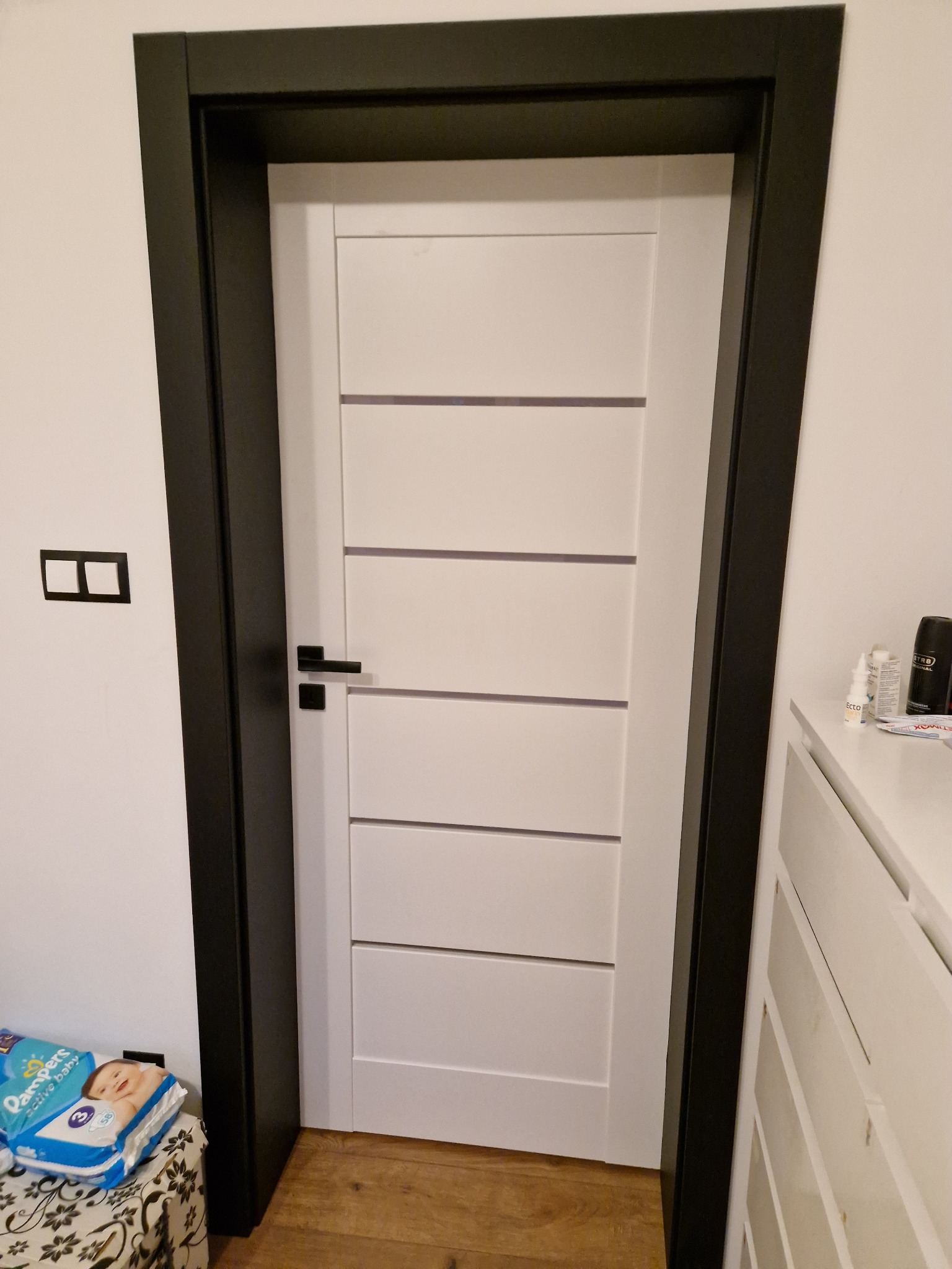 Białe drzwi wewnętrzne z poziomymi frezowaniami i czarną ościeżnicą, widoczny fragment pokoju z komodą i paczką pieluch.