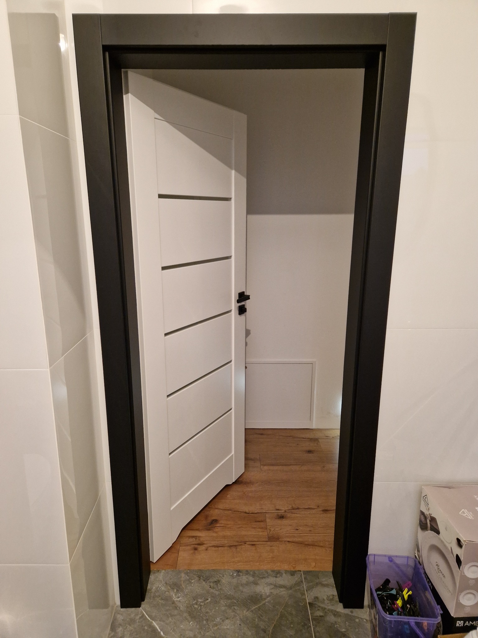 Białe drzwi wewnętrzne z czarną ościeżnicą, częściowo otwarte, ukazujące drewnianą podłogę w pomieszczeniu za nimi; widoczne białe płytki na ścianie i pudełko z akcesoriami w rogu.