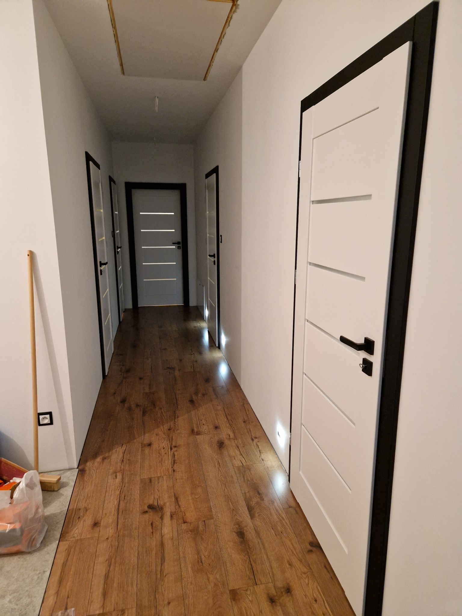 Korytarz z panelami podłogowymi w odcieniach brązu, białe drzwi z czarnymi ościeżnicami i klamkami, widoczne narzędzia i materiały budowlane przy ścianie.