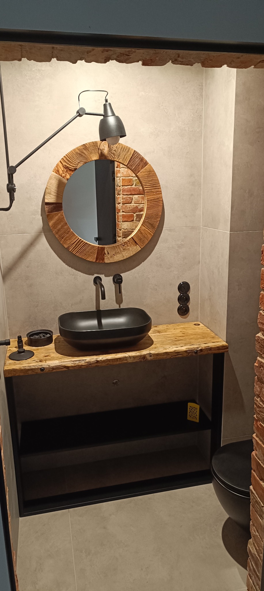 Nowoczesna łazienka z okrągłym lustrem w drewnianej ramie, czarną umywalką na drewnianym blacie i industrialną lampą.