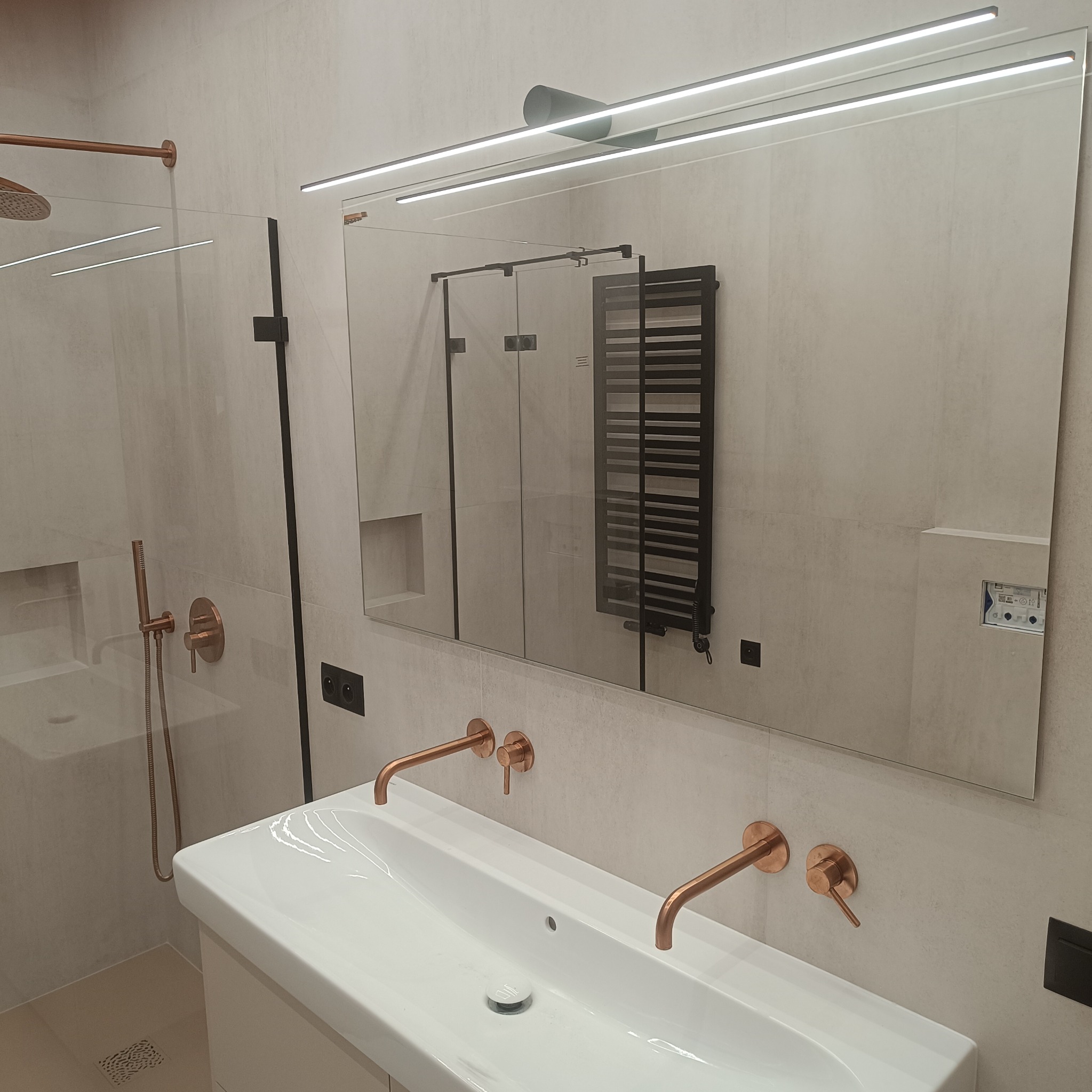 Nowoczesna łazienka z umywalką i bateriami w kolorze miedzi, kabina prysznicowa ze szklaną ścianką, lustro z podświetleniem LED i grzejnik drabinkowy widoczny w odbiciu.