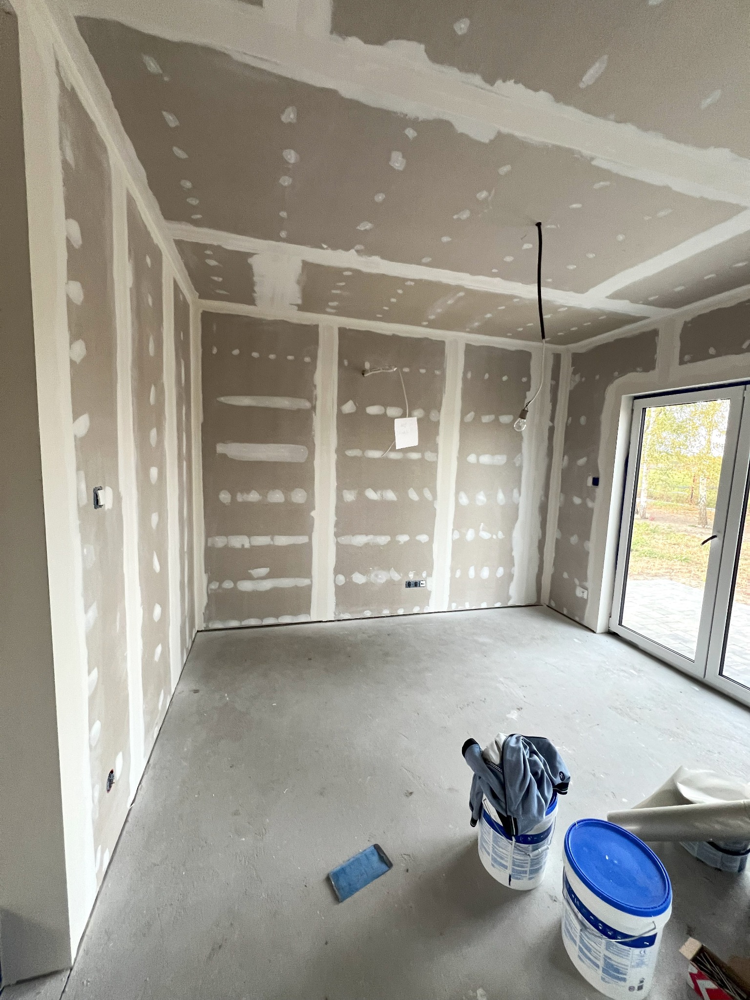 Wnętrze w trakcie wykańczania: ściany i sufit pokryte płytami, widoczne łączenia wypełnione masą, na podłodze dwa wiadra z niebieskimi pokrywkami i ubranie, okno z widokiem na zewnątrz.